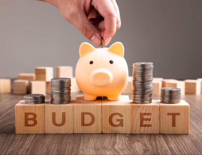 A hand dropping a coin into a piggy bank amid stacks of coins on top of the wooden blocks that spell "BUDGET."