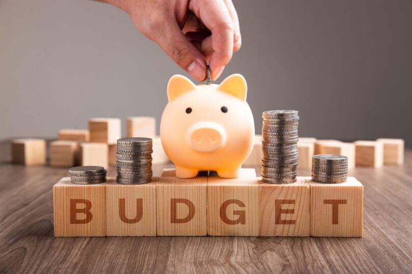 A hand dropping a coin into a piggy bank amid stacks of coins on top of the wooden blocks that spell "BUDGET."