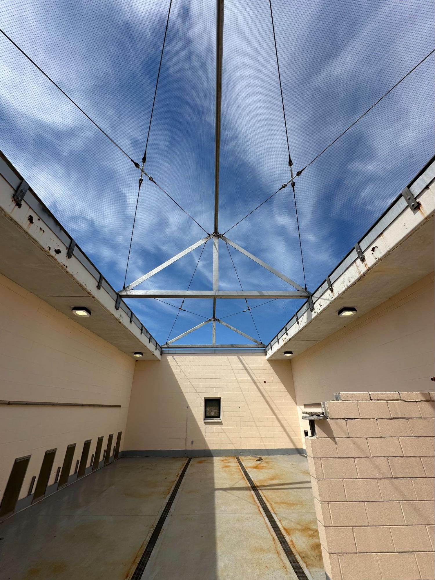 Open-air industrial courtyard seen from below, with a central metal frame and cables converging overhead against a blue sky with mesh roof.